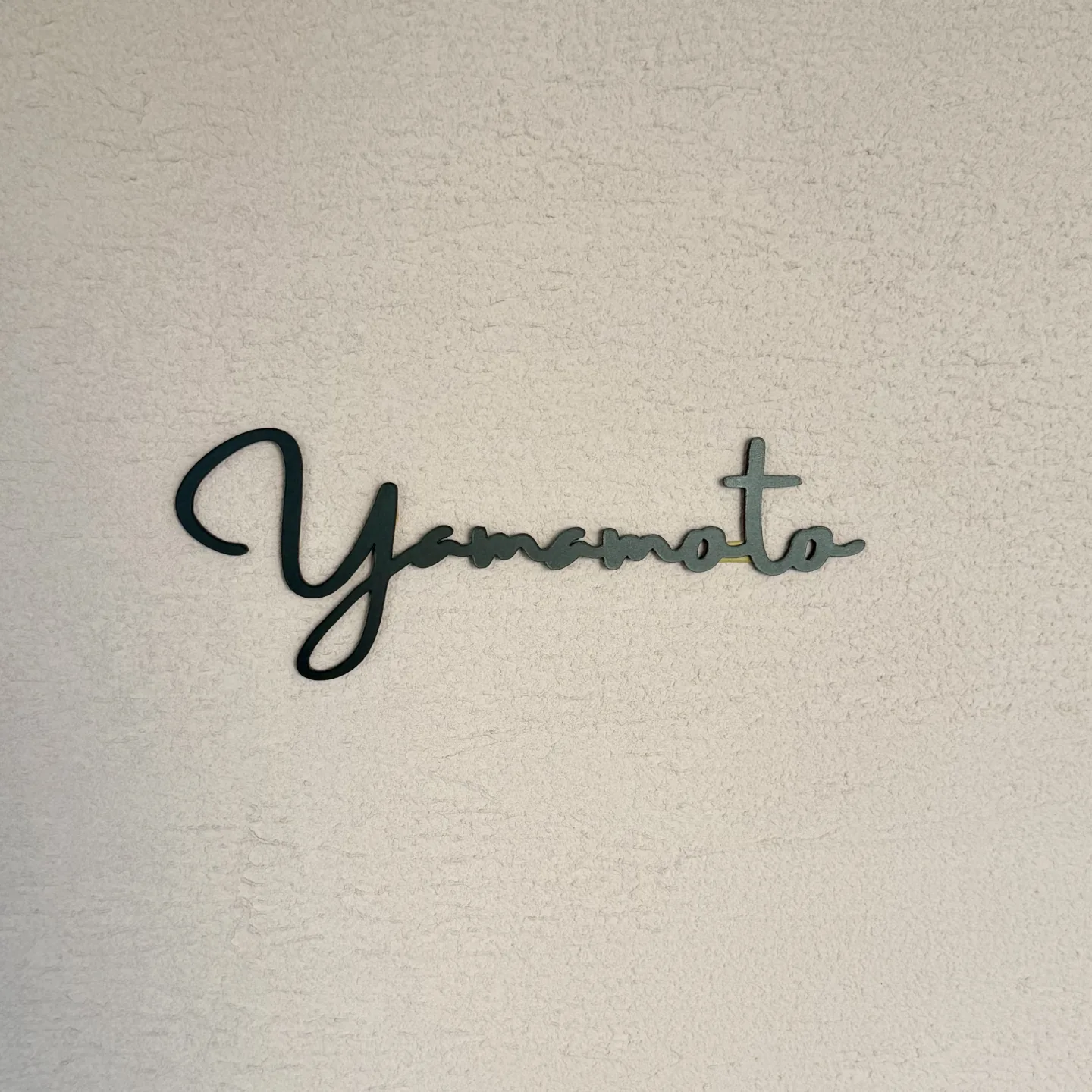 塗り壁を背景に撮影した筆記体 Yamamoto の切り文字表札